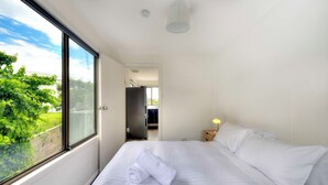 Une chambre d’hôtel avec une grande fenêtre, un lit avec du linge de maison blanc, une table de chevet avec une lampe, et une vue sur la verdure à l’extérieur.