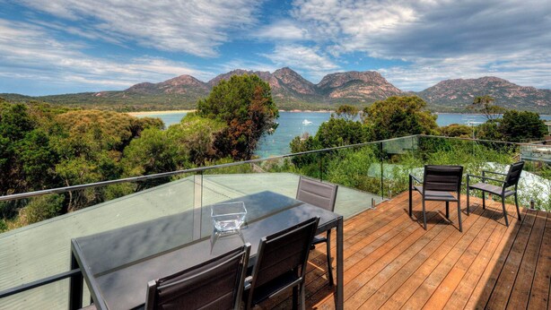 Une terrasse dotée d’une table et de chaises en verre, offrant une vue sur un paysage côtier avec des montagnes et une étendue d’eau.