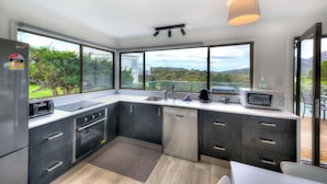 Une cuisine moderne équipée d’un lave-vaisselle, d’un four à micro-ondes et d’une cuisinière, de grandes fenêtres offrant une vue sur la verdure et un luminaire au plafond.
