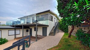 Une maison moderne à deux étages, dotée d’une façade en verre, d’une terrasse en bois et d’un chemin de gravier menant à un escalier.