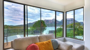 Une pièce avec un canapé beige, deux coussins et de grandes fenêtres donnant sur un lac et des montagnes.
