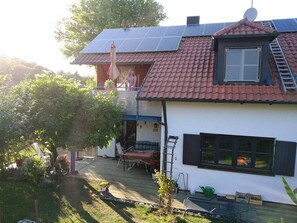 Une maison avec un toit recouvert de tuiles rouges, des panneaux solaires, un balcon et une terrasse aménagée avec du mobilier d’extérieur.