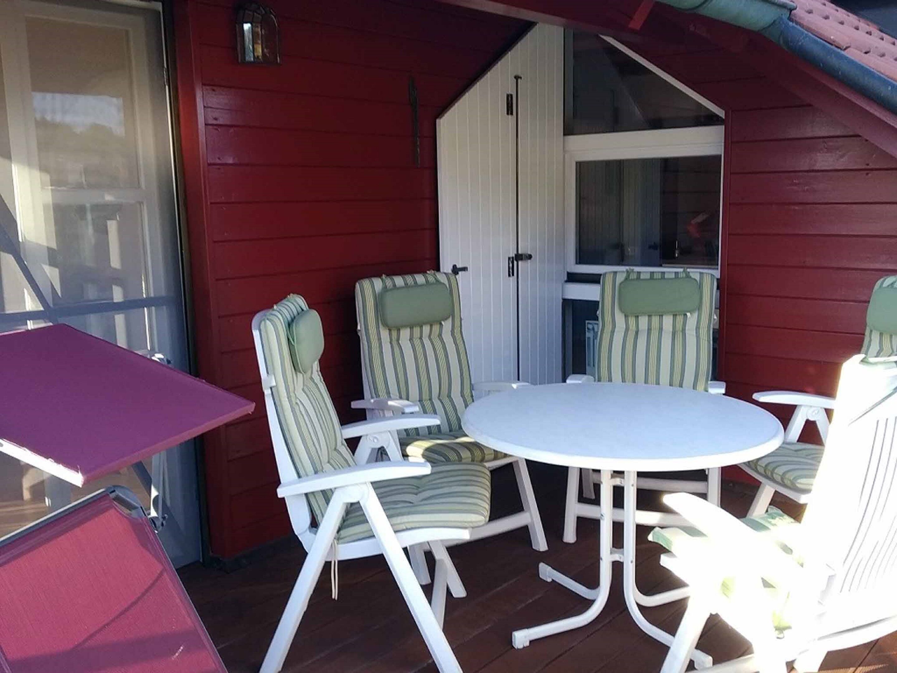 Ett takförsett utomhusområde med sittplatser, bestående av ett runt bord och fyra stolar, ett litet bord och en dörr som leder till en övre våning.