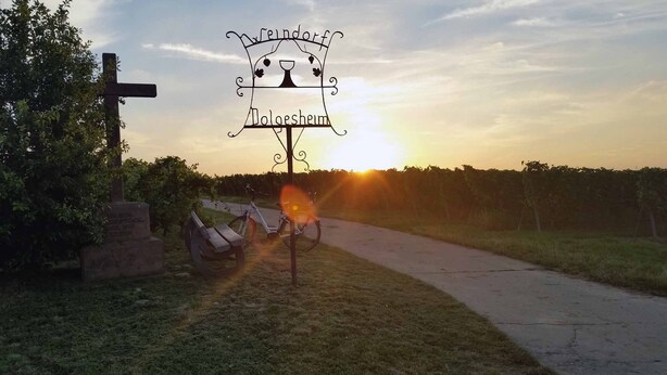 Taustalla auringonlasku, ja edessä kyltti, jossa lukee »Weindorf» ja »Loogesheim».