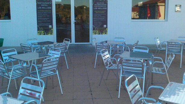 Un espace extérieur aménagé en terrasse, avec des tables et des chaises en métal, une entrée avec une porte vitrée, et deux affiches faisant la publicité d’un restaurant et d’une salle de réception.