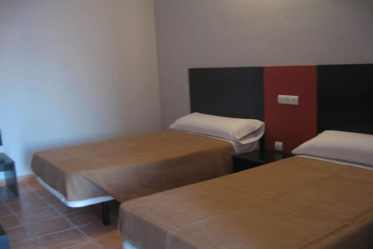 Habitación de hotel con dos camas individuales, cada una con una colcha marrón y almohadas blancas. La habitación tiene un diseño sencillo y funcional, con piso de baldosas y una pared lisa con un detalle rojo.