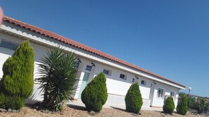 Un bâtiment avec un toit recouvert de tuiles rouges, des murs blancs et une allée gravillonnée. On y trouve trois arbres à feuilles persistantes verts et une plante ressemblant à un palmier à l’avant.