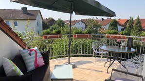 A balcony with a sofa, a small table, and a chair. There is a railing around the balcony, and a house is visible in the background. - Ferienwohnung Bellissima (Ering)