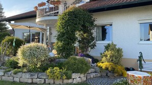 A house with a balcony, a chimney, and a garden with various plants and a stone wall. - Ferienwohnung Bellissima (Ering)