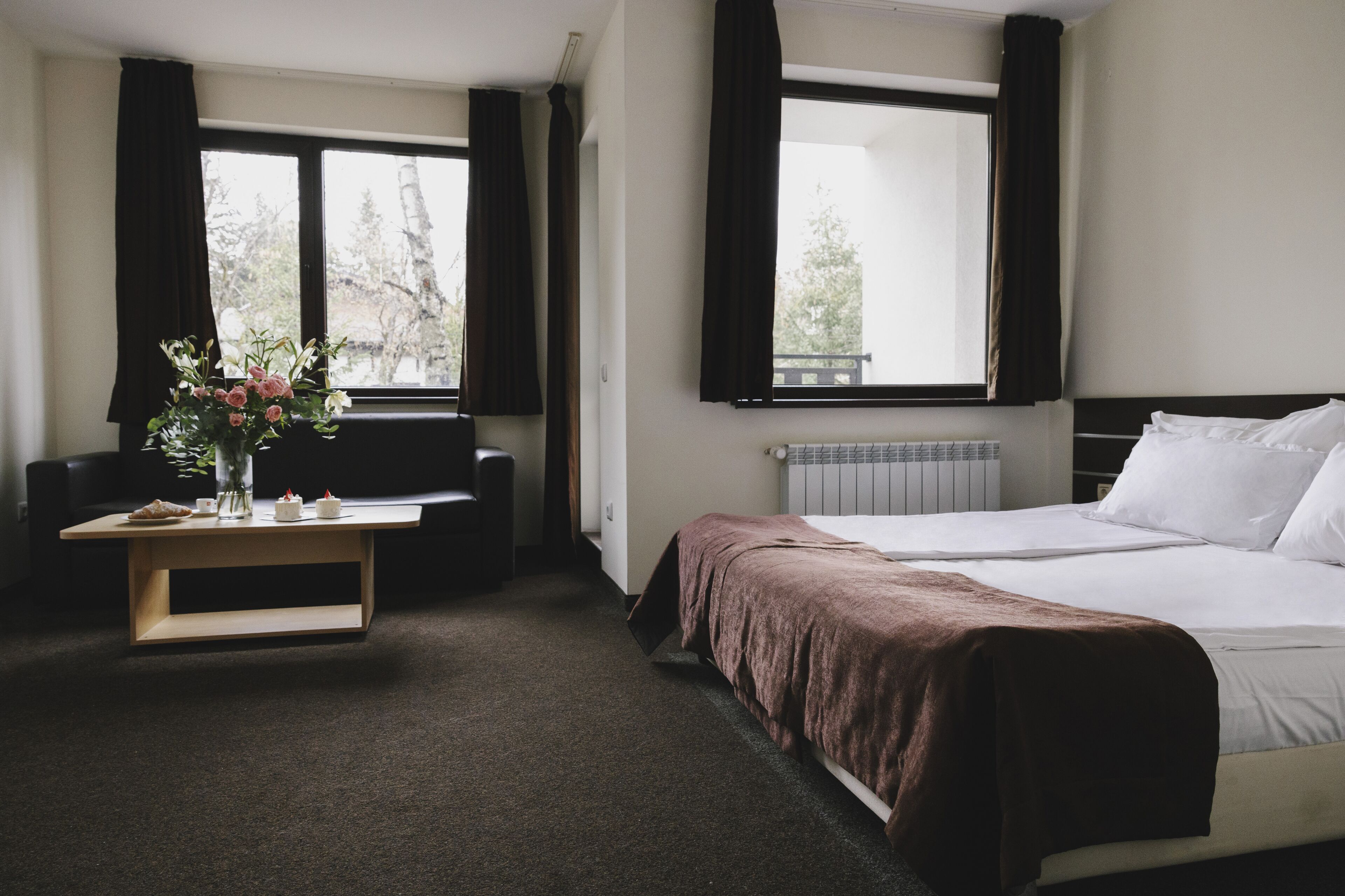 A hotel room with a bed, a desk with a vase of flowers, and a view of trees through the windows.