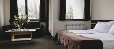 A hotel room with a bed, a desk with a vase of flowers, and a view of trees through the windows.