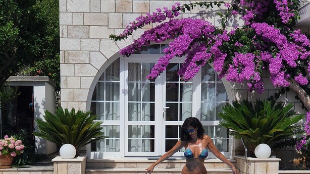 A woman in a bikini standing at the edge of a swimming pool with a stone building and purple flowers in the background. - Villa Ida (Dubrovnik)