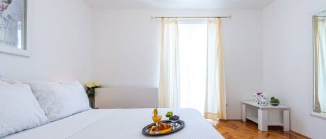 A hotel room with a large bed, a small bedside table with a tray of breakfast, a window with curtains, and a chandelier.