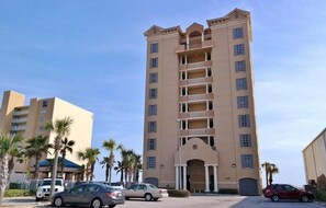 A multi-story hotel with a central entrance, balconies on each floor, and a parking area with several cars.