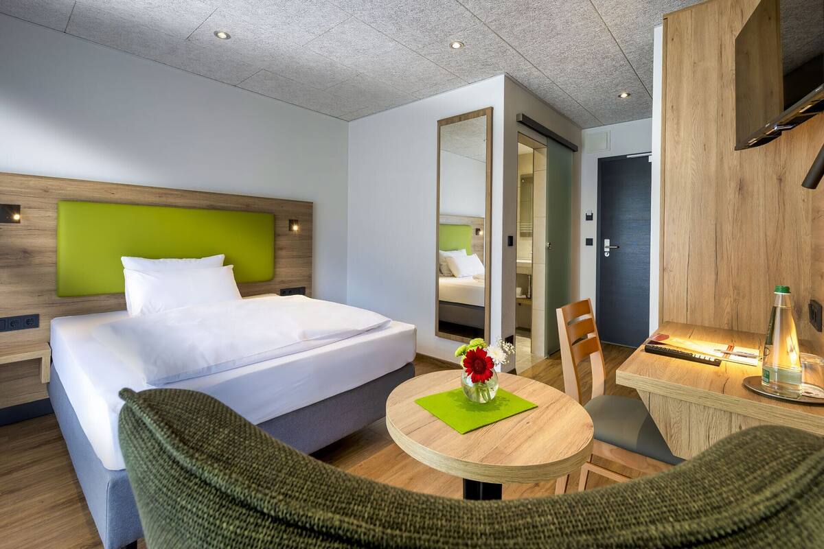 A modern hotel room with a bed, a round wooden table, a chair, a mirror, and a door.
