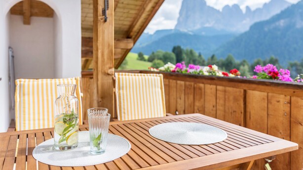 A wooden table with two chairs, a glass pitcher with a plant, and a clear glass. - Apartment “Lara Ruveda” with Mountain View, Private Terrace and Wi-Fi (St.Ulrich)