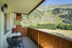 Un balcón con mesa y sillas, barandilla de madera y vistas a un paisaje montañoso.