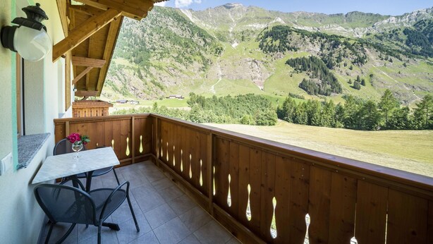 Un balcón con barandilla de madera, una mesa con sillas y vistas a montañas.