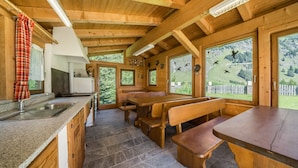 Interior de una cabaña de madera con cocina, comedor y ventanas amplias que dan a un paisaje montañoso.