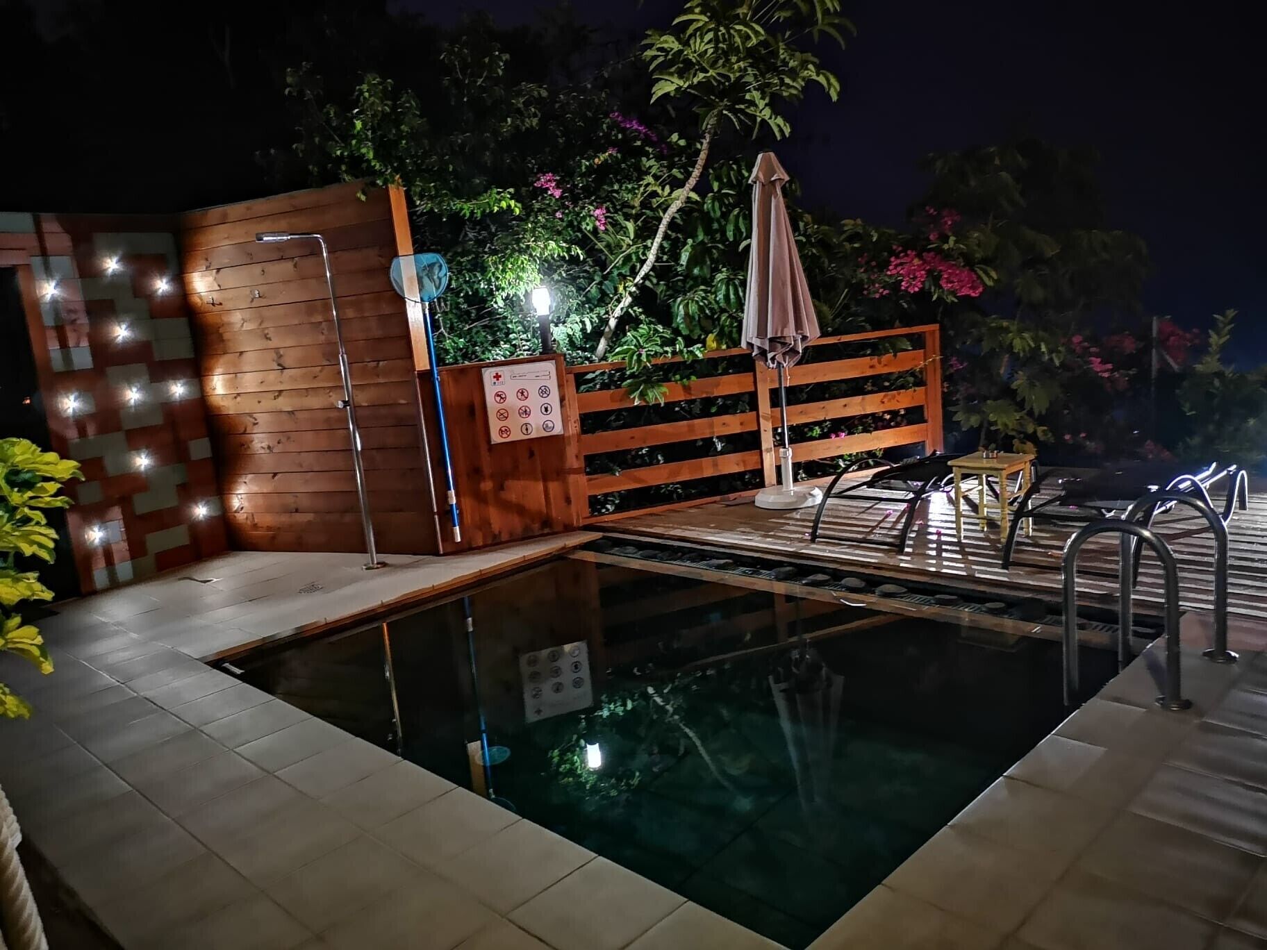 Una piscina con bordo in legno, recinzione metallica e un cartello con simboli.