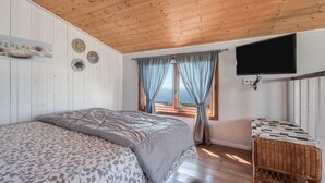 Una camera da letto con soffitto in legno, un letto con lenzuola decorate, una televisione montata a parete e vista attraverso una finestra con tende.