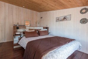 Een slaapkamer met een houten plafond, een bed met een bruine deken, een nachtkastje met een lamp en een spiegel aan de muur.