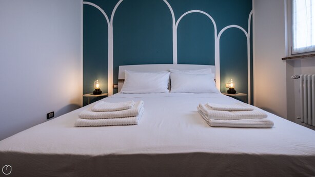 Una cama bien tendida con sábanas blancas y dos toallas dobladas encima. Hay dos mesitas de noche con lámparas a cada lado de la cama.