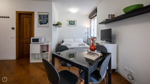 A compact hotel room with a bed, a desk with a chair, a microwave, a TV, and a shelf with books.