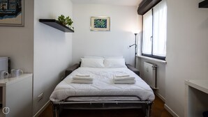 A small, clean room with a single bed, a shelf with a plant, a framed picture on the wall, and a window with curtains.
