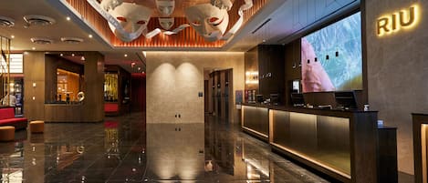 Un moderno vestíbulo de hotel con una recepción, una pantalla grande que muestra el nombre del hotel y un techo con máscaras teatrales.