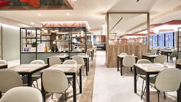Een modern restaurant met een lange toonbank, meerdere tafels en stoelen. Er zijn planken met decoratieve objecten en een uitzicht op de stad door grote ramen.