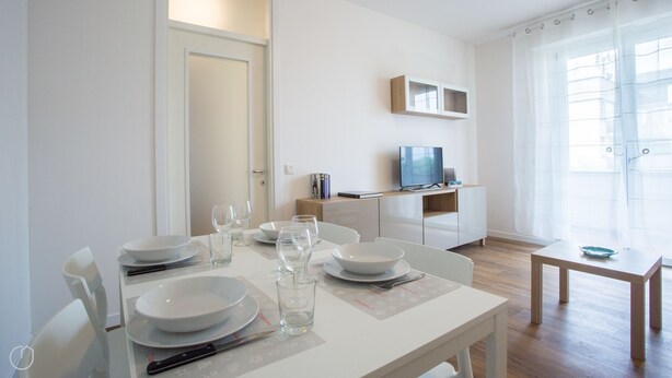 A dining area with a table set for four, a television on a stand, and a window with sheer curtains.