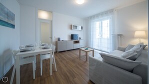 A modern living room with a sofa, a dining table with chairs, a TV, and a kitchen area with cabinets and a microwave.