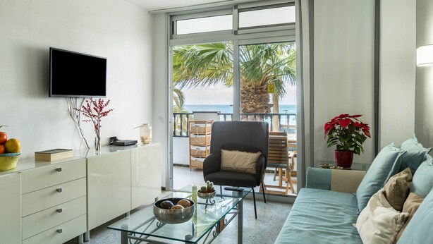 Una sala de estar con televisión, mesa de centro de vidrio, sofá, sillón y vista al mar a través de ventanas grandes.