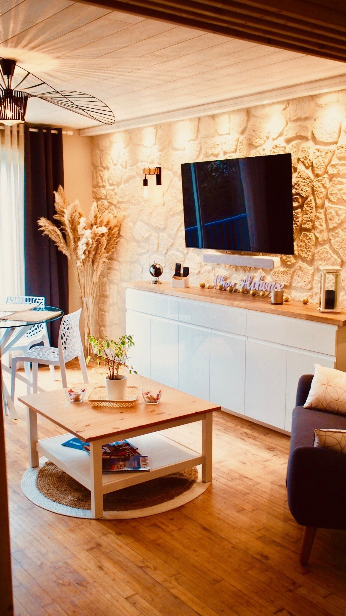 Een moderne woonkamer met een stenen muur, een flatscreen televisie, een houten salontafel, een bank en een eethoek met stoelen en een kleine tafel.