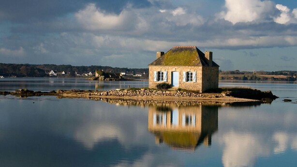 Une maison en pierre dotée d’une porte et de fenêtres, située sur une côte rocheuse, au bord d’une étendue d’eau calme dans laquelle se reflète la façade du bâtiment.