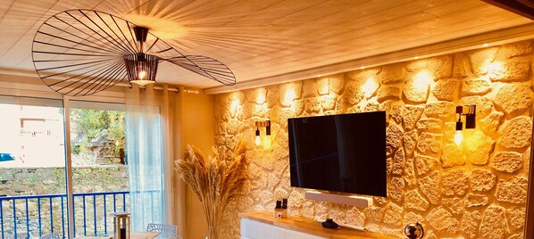 A room with a stone wall, a mounted TV, and a ceiling fan. - Chez Alphonse: cosy, renovated T3 - near the port (VANNES)