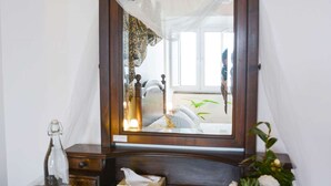 A bedroom with a wooden headboard, a bedside table with a bottle, glasses, and a tissue box. A mirror reflects a bed with pillows and a plant. - Pool suite with marble bath in exotic fruit orchard (Ribeira Grande)