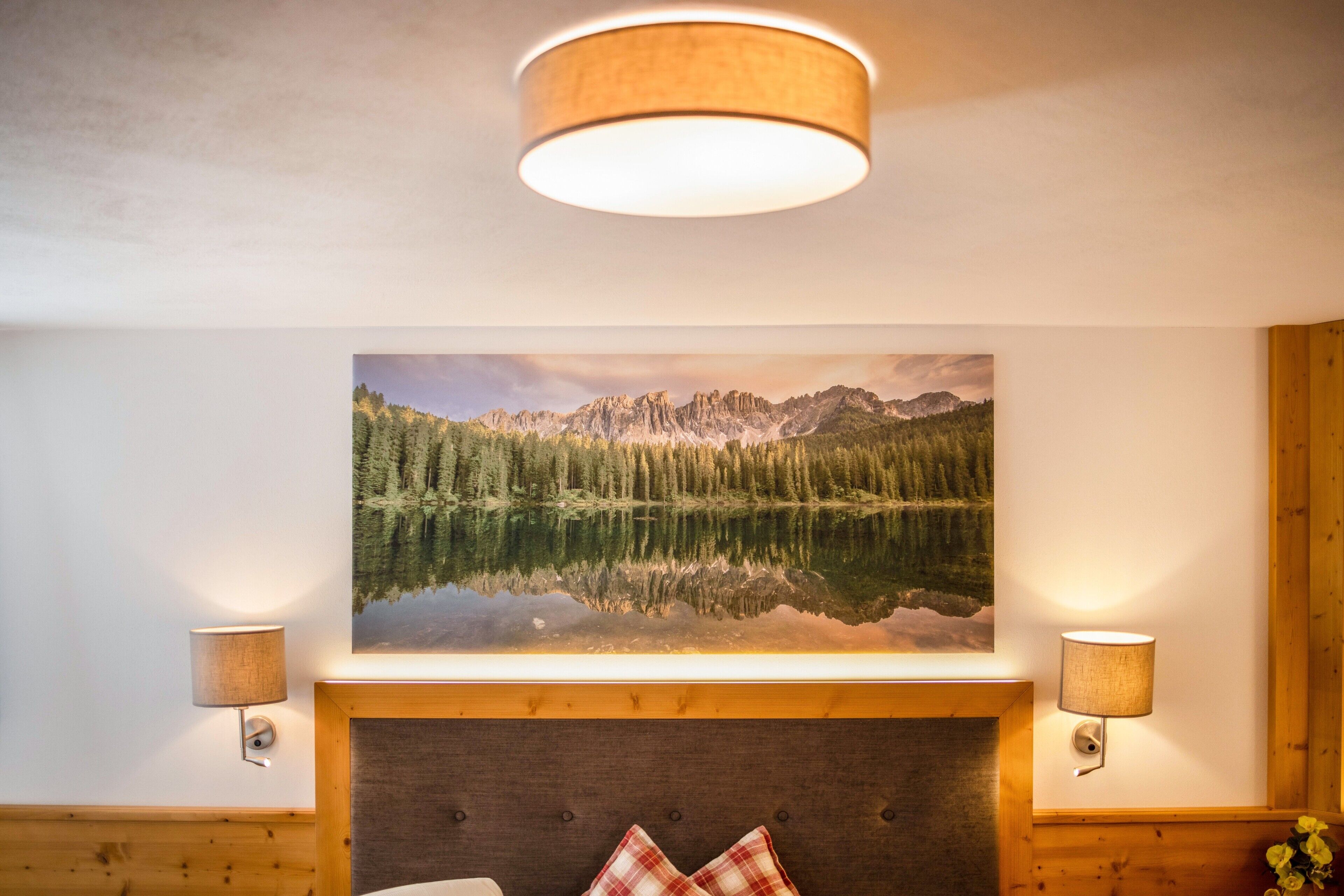 A bedroom with a large bed, a headboard, a wall-mounted painting of a mountain landscape, and two wall-mounted lamps.