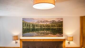 A bedroom with a large bed, a headboard, a wall-mounted painting of a mountain landscape, and two wall-mounted lamps.