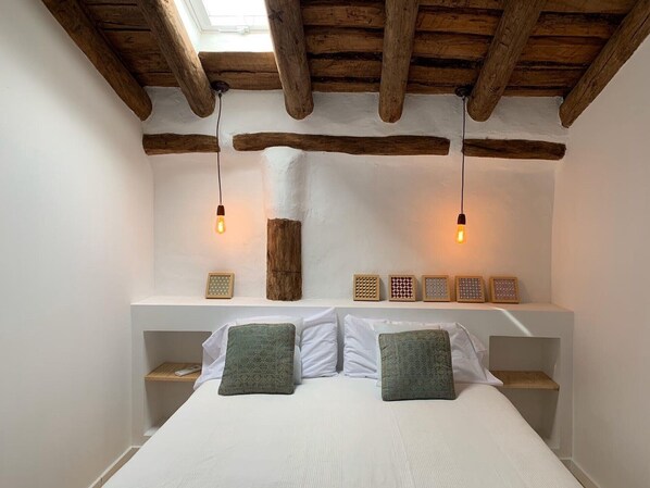 A bedroom with a wooden ceiling, a bed with white bedding, two bedside tables, and a shelf with decorative items.