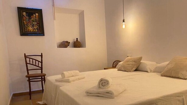 Een net opgemaakt bed met witte lakens, een stoel, een wandlamp en een ingelijste foto.