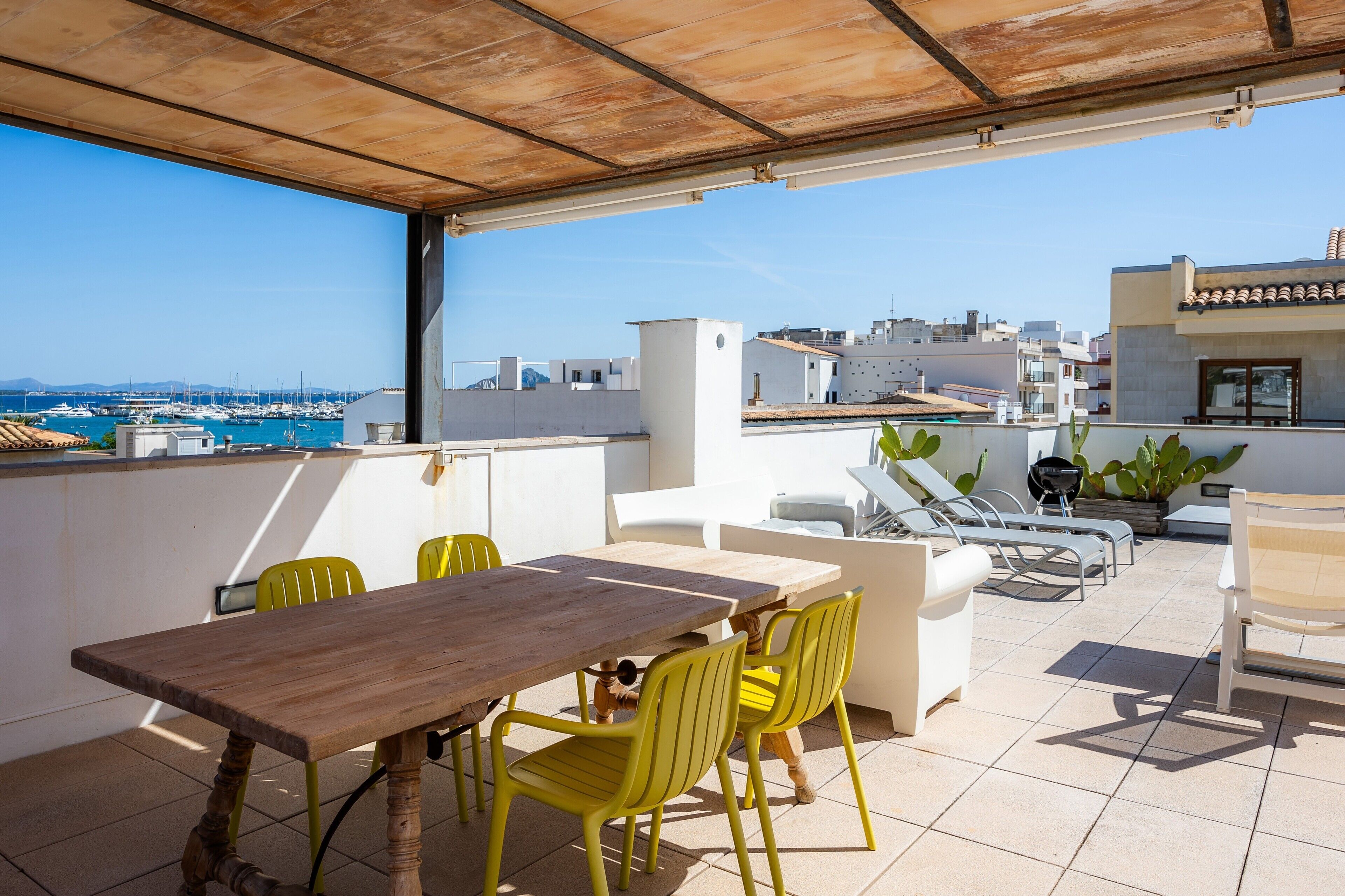 Une terrasse avec une table en bois et des chaises jaunes, donnant sur une marina et des immeubles.