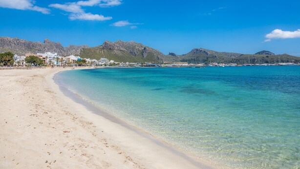 Une plage avec une eau turquoise et limpide, un rivage sablonneux et des bâtiments en arrière-plan.
