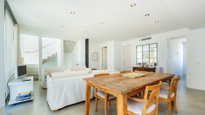 Un vaste espace de vie comprenant une table à manger en bois, des canapés blancs et un escalier menant à l’étage supérieur.