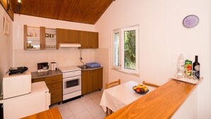 A compact kitchen with wooden cabinets, a white countertop, a dining table with a white tablecloth, and a window with a view of greenery. - Apartment Srđ  (Dubrovnik)