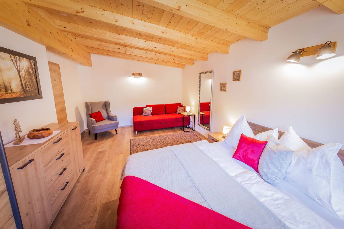 Ein Schlafzimmer mit Holzboden, einem Bett mit weißer und roter Bettwäsche, einem roten Sofa, einer hölzernen Kommode und einem Sessel.