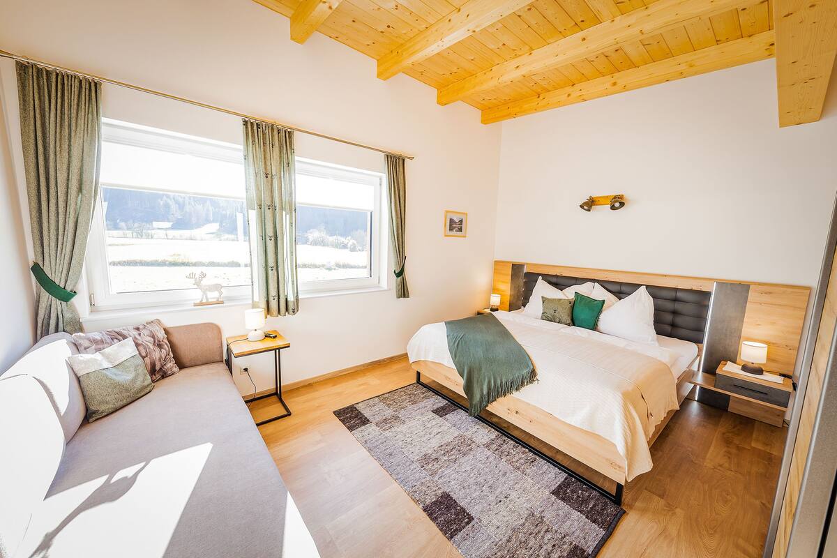 Ein Schlafzimmer mit Bett, Sofa, Nachttisch und einem Fenster mit Vorhängen.