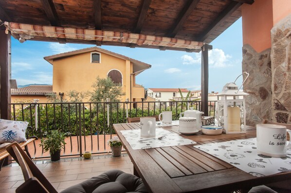 Ein Holzbalkon mit einem Tisch für zwei Personen, einem Blumentopf und Blick auf ein gelbes Gebäude mit rotem Dach.