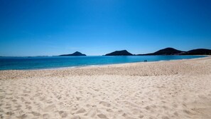 Une vaste plage de sable fin, bordée d’une eau d’un bleu limpide et dominée par des collines lointaines.